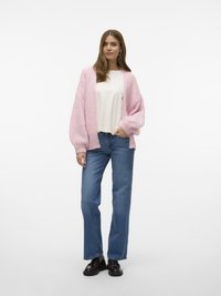 Donna che indossa un cardigan chunky rosa chiaro sopra una maglietta bianca, jeans a gamba dritta blu e mocassini neri, posando su uno sfondo bianco.