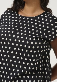 Haut noir avec un col rond et des manches courtes, arborant un motif à pois blancs. Le tissu semble lisse et léger.