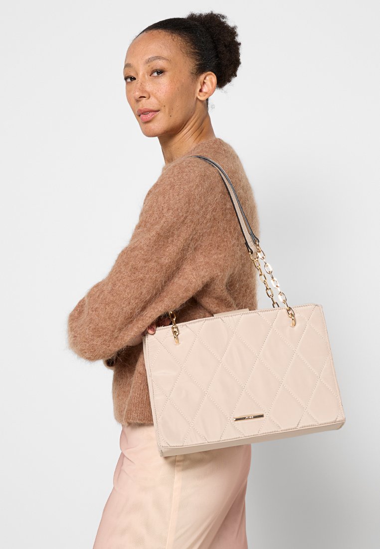 Sac à main beige matelassé avec quincaillerie dorée et bandoulière en chaîne, présentant une texture lisse et une forme rectangulaire. Accent décoratif à l'avant.
