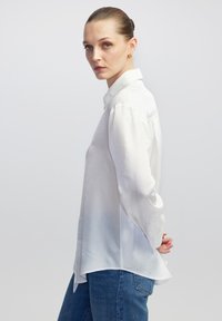 Biała koszula z długim rękawem i kołnierzykiem, o gładkiej fakturze i bocznych rozcięciach, w połączeniu z niebieskimi dżinsami, prezentowana w widoku bocznym.