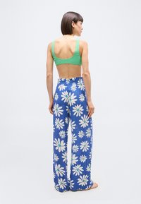 Γυναίκα όρθια, ξυπόλητη και με την πλάτη στραμμένη, φοράει πράσινο κοντό τοπ και μπλε παντελόνια με φαρδιά μπατζάκια και λευκό μοτίβο με λουλούδια μαργαρίτας.