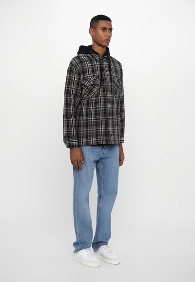 Veste en flanelle à carreaux noir et blanc avec capuche, deux poches poitrine, assortie à un jean bleu clair et des baskets blanches.