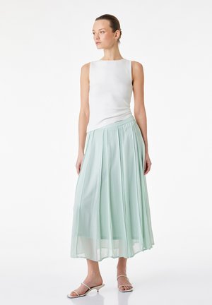 Femme debout, portant un top blanc sans manches, une jupe midi plissée vert menthe clair et des sandales blanches à talons, regardant sur le côté.