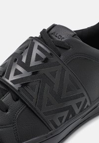 Svart idrottssko med en texturerad läderöverkonstruktion, ett geometriskt mönstrat rem och släta accenter för en modern design.