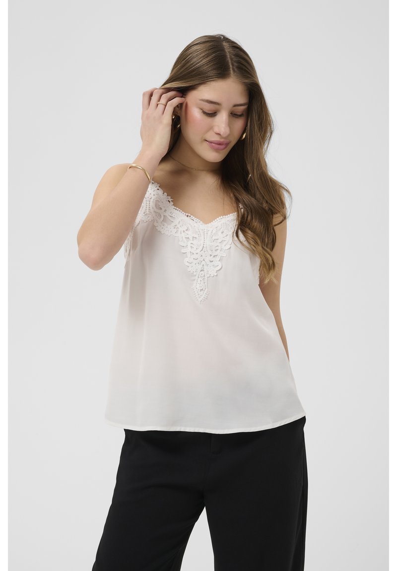 Jonge vrouw met lang bruin haar, gekleed in een mouwloos wit topje met kanten afwerking en zwarte broek, die haar haar aanraakt, tegen een effen achtergrond.