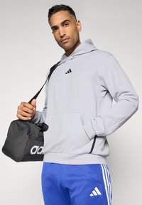 Felpa grigia chiara con dettagli neri e logo, tasca frontale, indossata con pantaloni jogger blu con strisce bianche. Borsa a tracolla nera con logo.