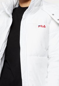 Hvid polstret jakke med quiltet design og lynlåslukning. Har en FILA-logo i rød og blå på brystet. Tekstureret materiale.