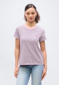 Νεαρή γυναίκα με σγουρά μαλλιά και σκουλαρίκια κρίκους φοράει κοντομάνικη ριγέ μπλούζα σε ροζ, λευκό, κόκκινο και ναυτικό μπλε και ανοιχτόχρωμο τζιν παντελόνι.