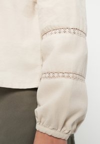 Kermanvalkoinen pusero, jossa on pitkät pussihihat ja pitsikoriste lähellä hihansuutä. Kangas on pehmeää ja siinä on hienovarainen ribbineulos.