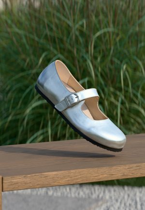 Silberner Mary Jane-Schuh mit Schnallenriemen, schwarze Sohle, liegend auf einer Holzoberfläche mit grünem Gras im Hintergrund.