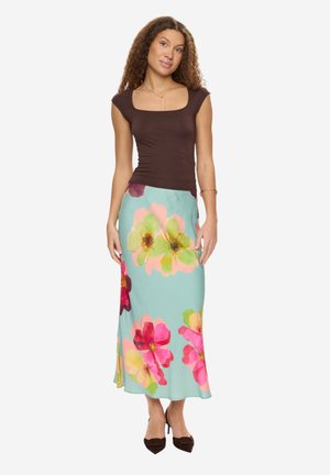 Nainen seisoo, pukeutunut istuvaan ruskeaan toppiin ja vaaleansiniseen hameeseen, jossa on suuret vaaleanpunaiset, keltaiset ja vihreät kukkakuviot, yhdistettynä ruskeisiin korkokenkiin.