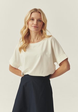 Weiße Bluse mit kurzen Ärmeln, rundem Ausschnitt und hochgekrempelten Manschetten, kombiniert mit einem dunkelblauen Rock, der eine glatte Textur und eine lockere Passform aufweist.