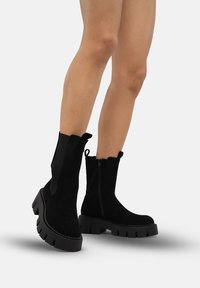 Black ankle boots made of suede with a chunky sole, featuring a zipper and elastic side panels. The design has a scalloped top edge.