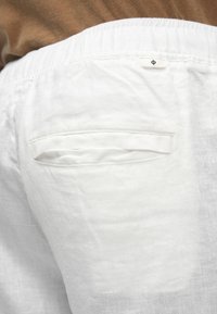 Hvide linnedbukser med elastik i taljen, der har en enkelt baglomme og et lille mærke på taljen. Blød tekstur.