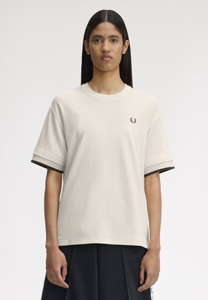 Donna con i capelli lunghi e scuri che indossa una maglietta beige a maniche corte con un piccolo emblema rosso e una gonna scura, in piedi davanti a uno sfondo semplice.