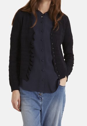 Marineblauer Strickcardigan mit strukturierten Mustern, Rüschendetails an der Vorderseite und Knopfverschluss. Über einer passenden marineblauen Bluse getragen.