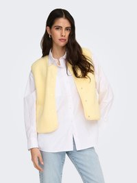 Geelt fleecesvest met drukknoopsluiting over een wit overhemd met knopen, gecombineerd met lichtblauwe spijkerbroek. Glad oppervlak, comfortabele pasvorm.