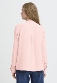 Camicia rosa a maniche lunghe con yoke posteriore e dettagli a pieghe, abbinata a jeans di denim blu scuro. Tessuto liscio, polsino con bottoni, design casual.
