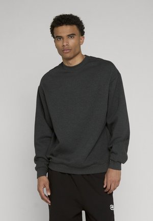 Junger Mann in einem dunkelgrauen Langarm-Sweatshirt und schwarzen Hosen, stehend vor einem einfarbigen hellgrauen Hintergrund.