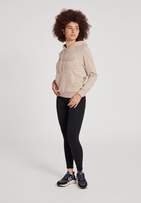 Haut à capuche beige avec une poche avant et un cordon de serrage, arborant un logo en relief. Accompagné de leggings noirs et de chaussures de course.