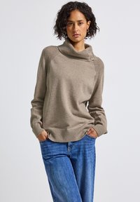 Beige gebreide trui met een hoge kraag, voorzien van een gestructureerd schouderdetail en drie knopen aan één kant; gedragen met blauwe denim jeans.