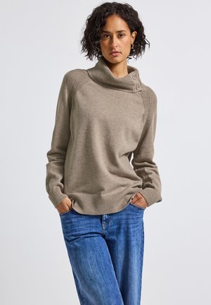 Beige strikket sweater med høj krave, med teksturerede skulderdetaljer og tre knapper på den ene side; båret med blå denim jeans.