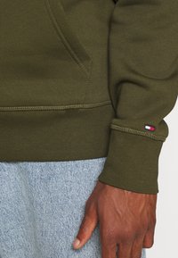 Acercamiento de una persona que lleva puesto un sudadera verde oliva con detalle de bolsillo cosido y un pequeño logotipo de bandera en el puño, combinada con unos jeans de mezclilla azul claro.