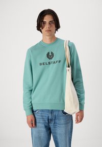 Türkiissinine kapuutsiga sviiter musta "BELSTAFF" logo, ümmargune kaelus, pikad varrukad. Kombineeritud helesiniste teksade ja kreemikat värvi kottiga.