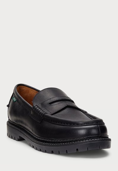 Mocasin de cuero negro con un diseño cosido, suela plana con tracción y una banda decorativa a lo largo del empeine. El interior es de color marrón.