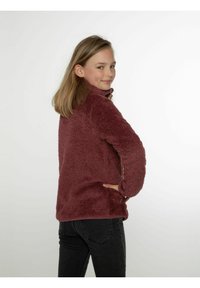 Pullover in pile bordeaux con colletto alto, texture morbida, tasche laterali e maniche lunghe; abbinato a jeans scuri.