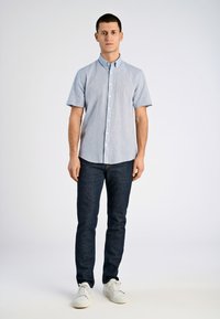 Camicia blu chiaro a maniche corte con bottoni, jeans blu scuro, sneakers bianche, in piedi su uno sfondo semplice. Tessuto morbido, design a vestibilità rilassata.