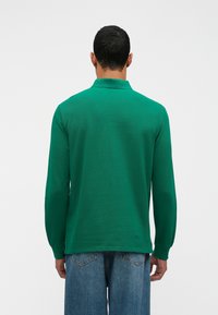 Zelená polokošeľa s dlhým rukávom a golierom, vyrobená z textúrovaného materiálu, s rovnými okrajmi a bočnými rozparkami, kombinovaná so svetlomodrými džínsami.