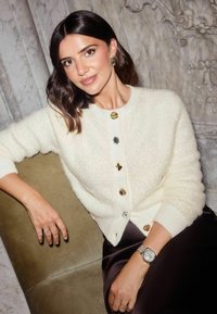 Slonovinový chlpatý kardigán so zlatými, striebornými a čiernymi dekoratívnymi gombíkmi, nosený cez tmavú saténovú sukňu. Ruka modelky spočíva na zelenej pohovke.