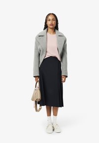 Abrigo de lana gris con solapas anchas, camiseta rosa claro, falda azul marino con pliegues, calcetines blancos, zapatillas crema, sosteniendo un bolso beige con acentos.