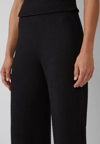 Schwarze, strukturierte Weitbein-Hosen mit einem krausigen Muster, ausgestattet mit einem eng anliegenden Bund. Nahaufnahme, die die Oberfläche des Stoffes und die Details zeigt.