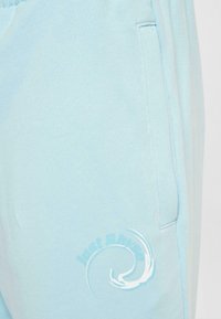 Light blue sweatpants made of soft fabric, featuring a side pocket and a white wave graphic with the text "just ride." Textured surface.