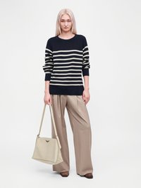Femme aux cheveux blonds raides portant un pull à rayures noir et blanc et un pantalon large beige, tenant un grand sac à main crème.