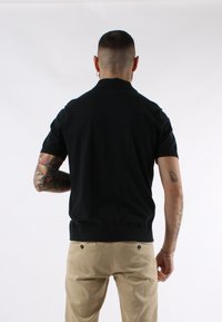 Polo shirt nero a maniche corte con una texture morbida, dotato di colletto classico e orlo a coste, indossato con pantaloni beige chiaro.