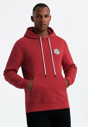 Röd hoodie med ribbade mudd och nederkant, dragsko-hette och framficka. Har en rund logotypklistermärke på bröstet. Slät textur.