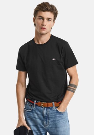 Čierne bavlnené tričko s okrúhlym výstrihom, priliehavý strih a malé logo na hrudi, v kombinácii s modrými džínsami a hnedým opaskom.