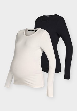 Schwangere Frau trägt beige Oberteil und schwarze Hose, hält eine große schwarze Clutch und steht vor einem schlichten hellen Hintergrund.