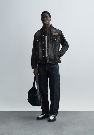 Μαύρο τζιν μπουφάν με κίτρινη κέντημα, συνδυασμένο με σκούρα τζιν και μαύρα παπούτσια με λευκές λεπτομέρειες. Αξεσουάρ περιλαμβάνει μια μαύρη τσάντα.