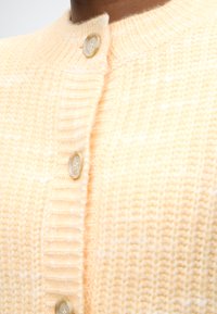 Πλεκτή ζακέτα από μαλακό, ανοιχτό κίτρινο νήμα με ριμπ γιακά. Διαθέτει δύο κουμπιά με διακριτική λάμψη, προσθέτοντας υφή στη λεπτομέρεια.