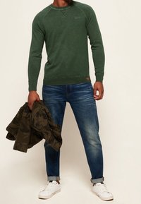 Grüner Baumwollpullover mit Raglanärmel, kombiniert mit blauen Jeans und einer Militärjacke. Weiße Sneaker runden den Look ab.