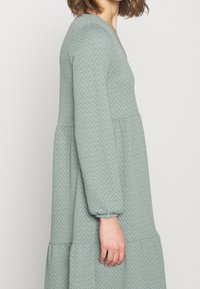 Robe à manches longues, à volants, en tissu vert texturé avec un motif en chevron. Présente un décolleté en V et des manches froncées aux poignets.