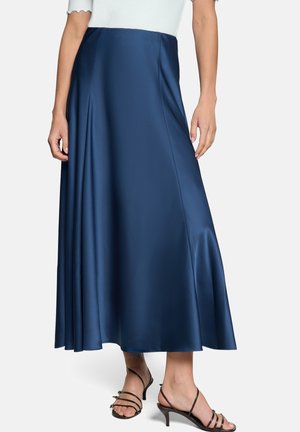 Frau trägt einen langen, marineblauen Satinsrock mit schwarzen, mit Riemchen versehenen Sandaletten mit Absatz vor weißem Hintergrund.