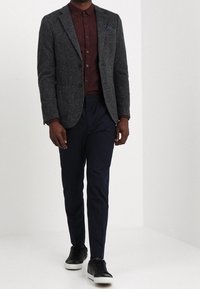 Uomo che indossa un blazer grigio scuro, una camicia bordeaux con bottoni, pantaloni blu navy e scarpe da ginnastica nere con suole bianche, che cammina in uno spazio bianco uniforme.