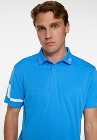 Blå polo skjorta i slät tyg, med knäppkrage, korta ärmar och vita detaljer på ärmarna samt en logotyp.