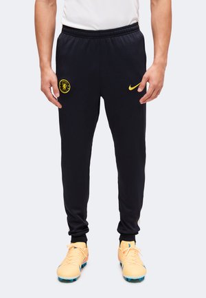 Mand iført sorte sportspants med gule Chelsea FC- og Nike-logoer, kombineret med lyse orange fodboldstøvler med blå knopper.