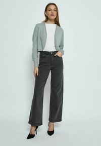 Cardigan grigio corto sopra una maglietta bianca, abbinato a jeans larghi neri a vita alta e tacchi a punta neri. Texture morbide e design semplice.